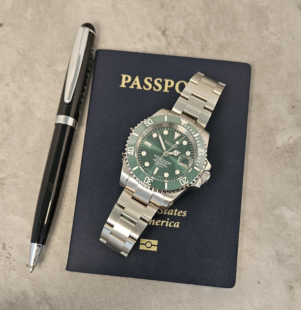 A custom mod Seiko watch with a green dial and green ceramic bezel, a silver stainless steel bracelet, displayed on top of a passport with a pen next to it.