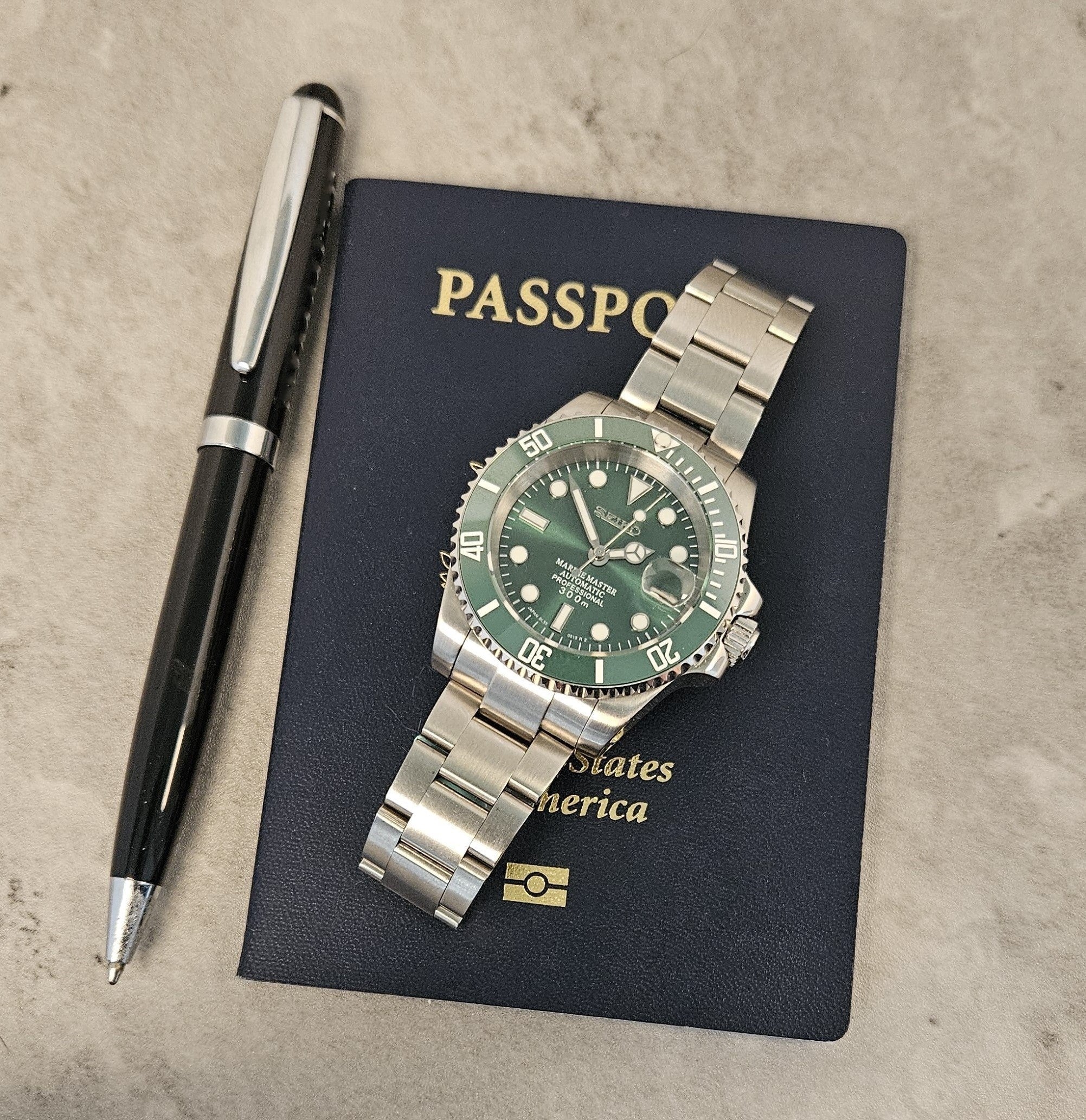 A custom mod Seiko watch with a green dial and green ceramic bezel, a silver stainless steel bracelet, displayed on top of a passport with a pen next to it.