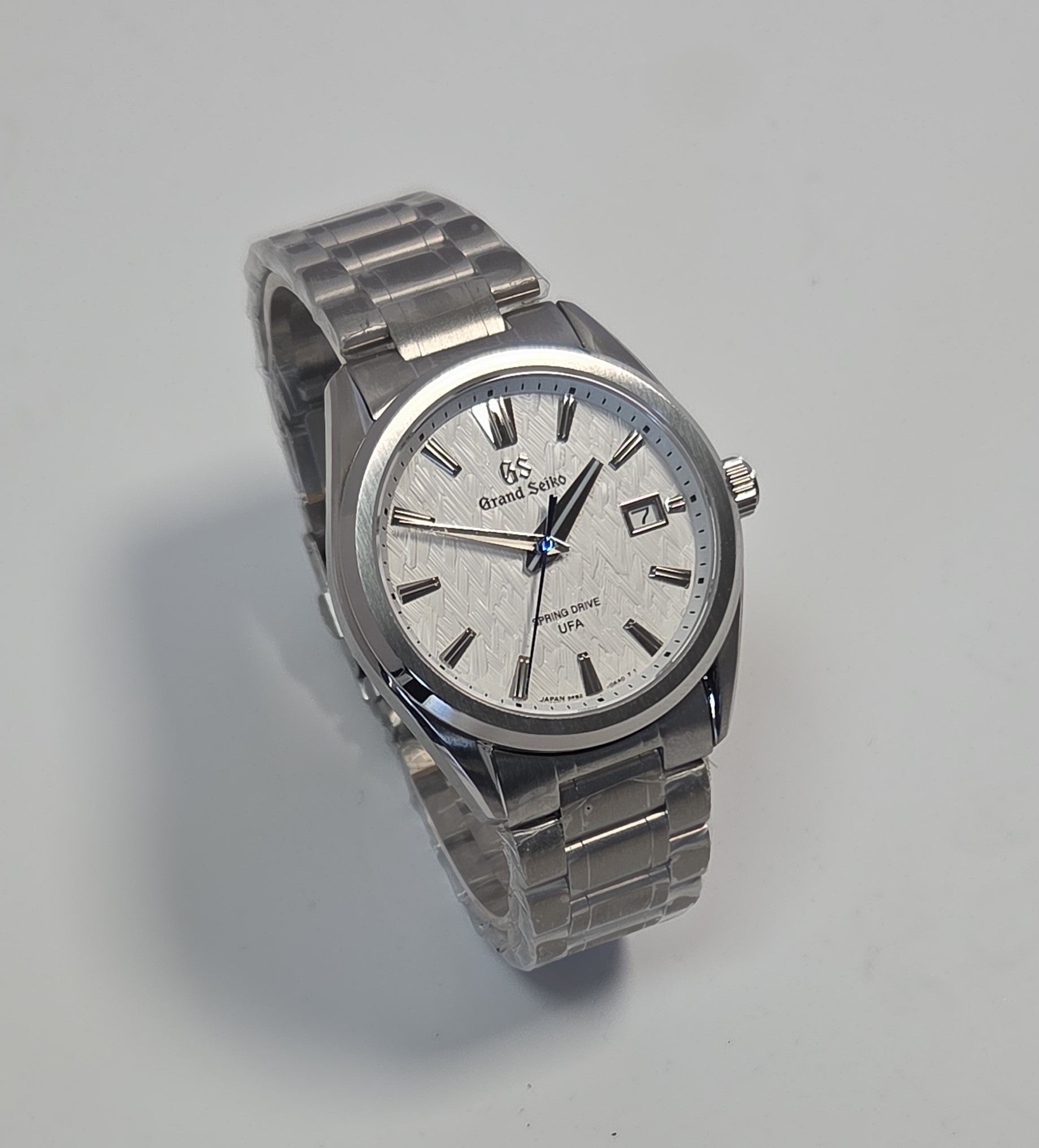 A silver stainless steel wrist watch with a textured white dial, featuring a date window, silver-tone hour markers, and a matching metal link bracelet, photographed on a light gray background.