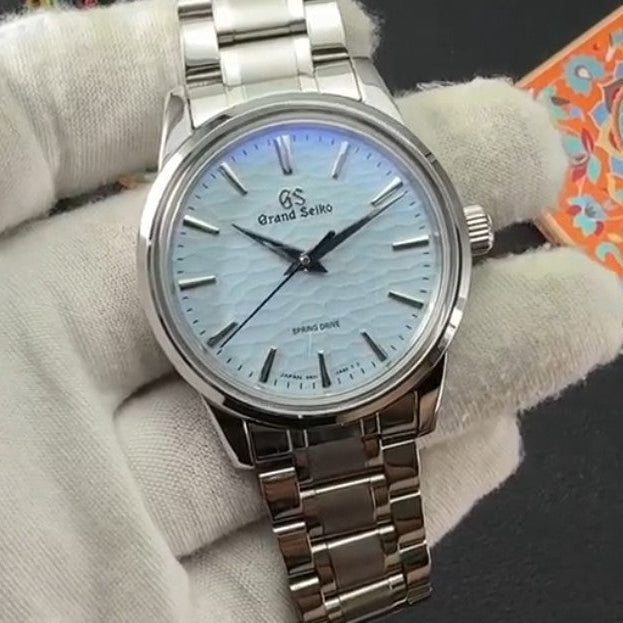 A stainless steel wristwatch with a silver-toned metal link bracelet and a light blue textured dial, held by a white-gloved hand against a dark background.