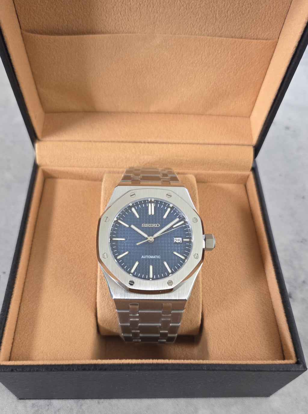 Silver watch with blue face in an open brown box on a gray surface
